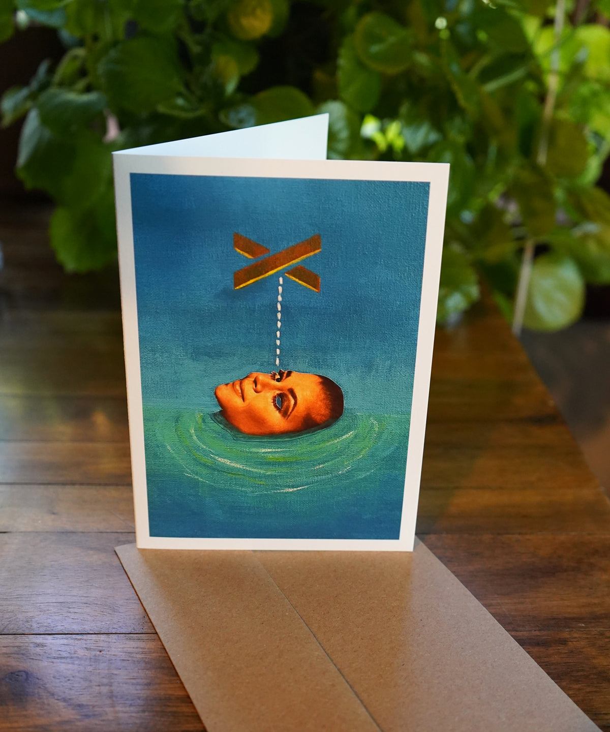 Greeting card with a face in water and a cross above it on a wooden surface with plants in the background.