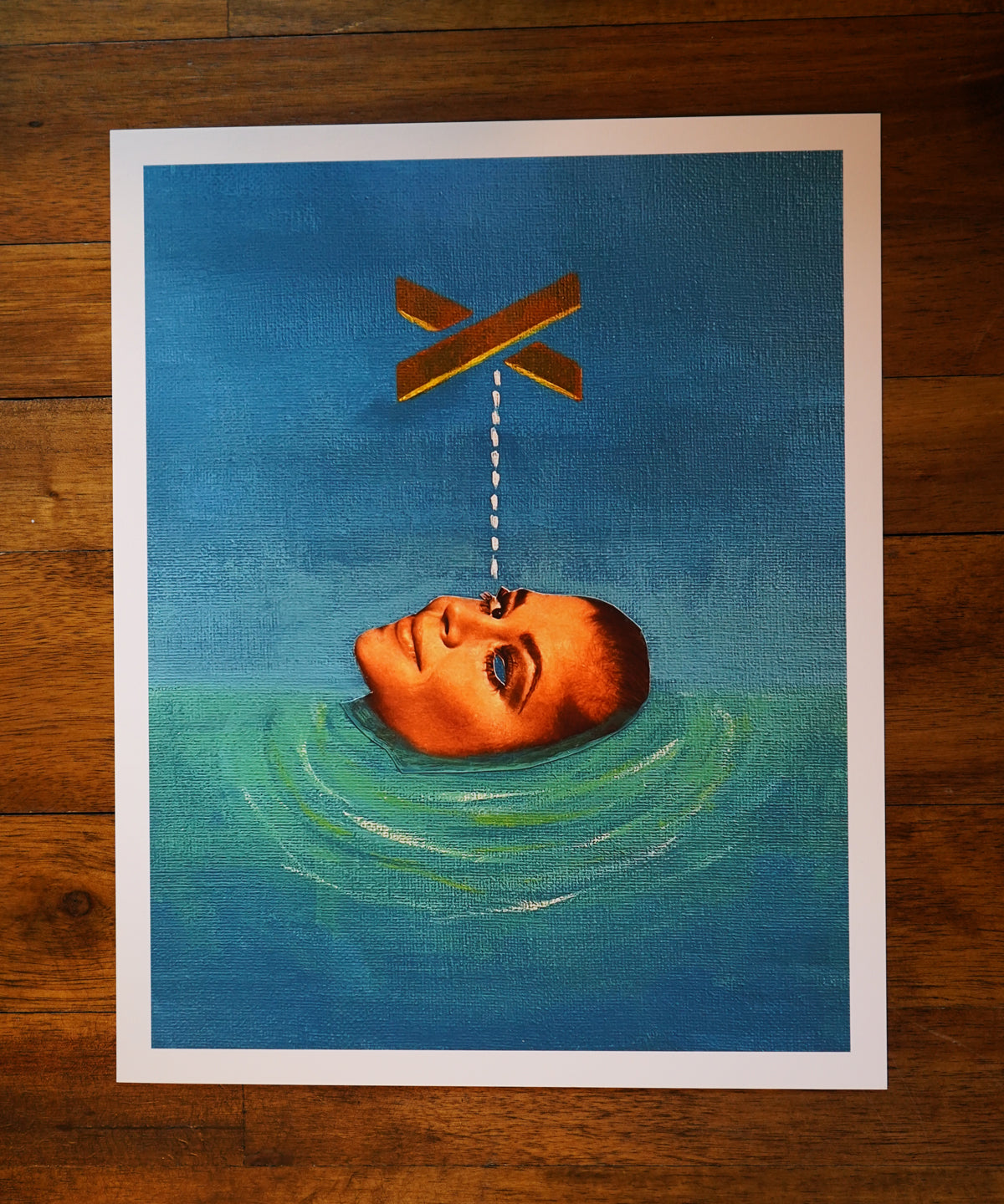 Art print of a head floating in water with an X above it on a wooden surface.