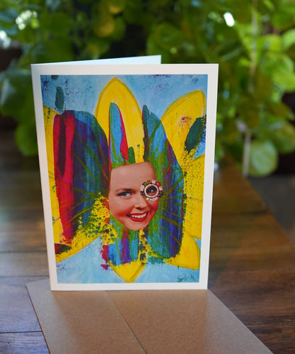 Colorful greeting card with a stylized face and flower petals on a wooden surface.