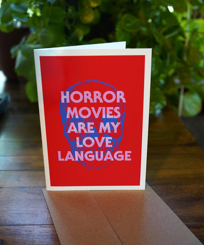 Card with 'Horror movies are my love language' text on a red background, placed on a wooden surface with plants in the background.